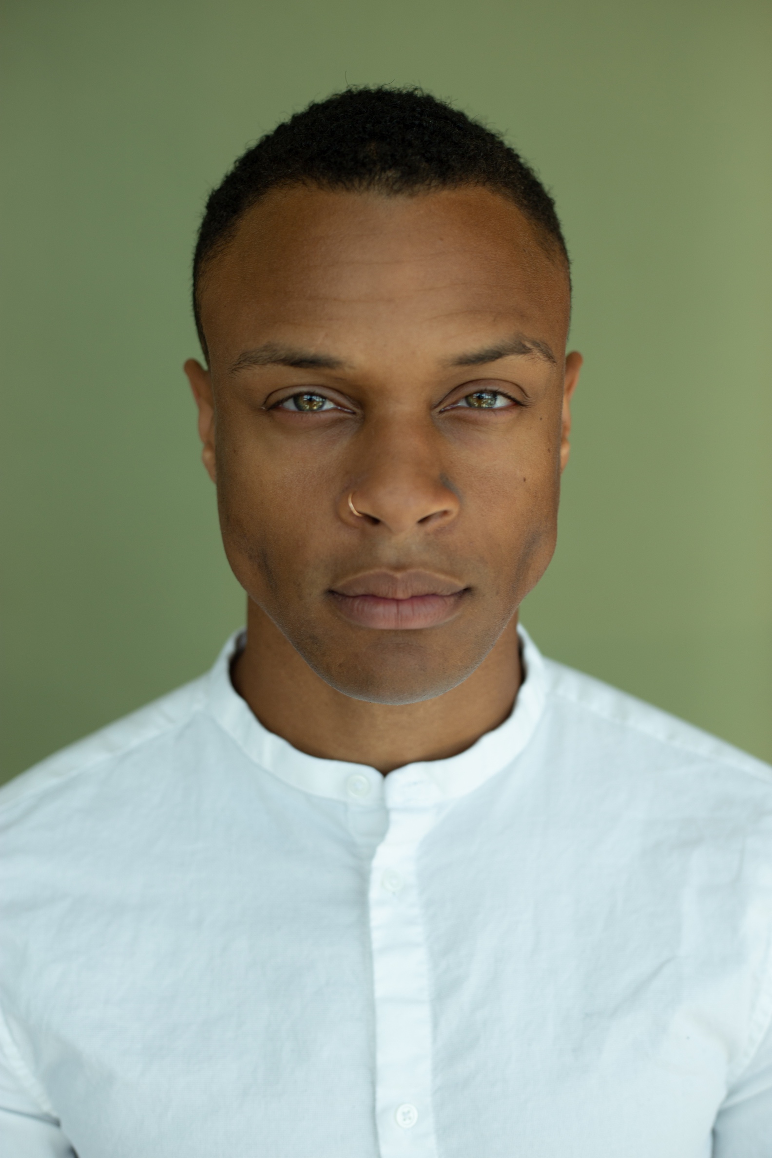 headshot of Joe, a Black man with caramel skin, short black hair, and hazel eyes, wearing a white shirt, and a gold nose ring 