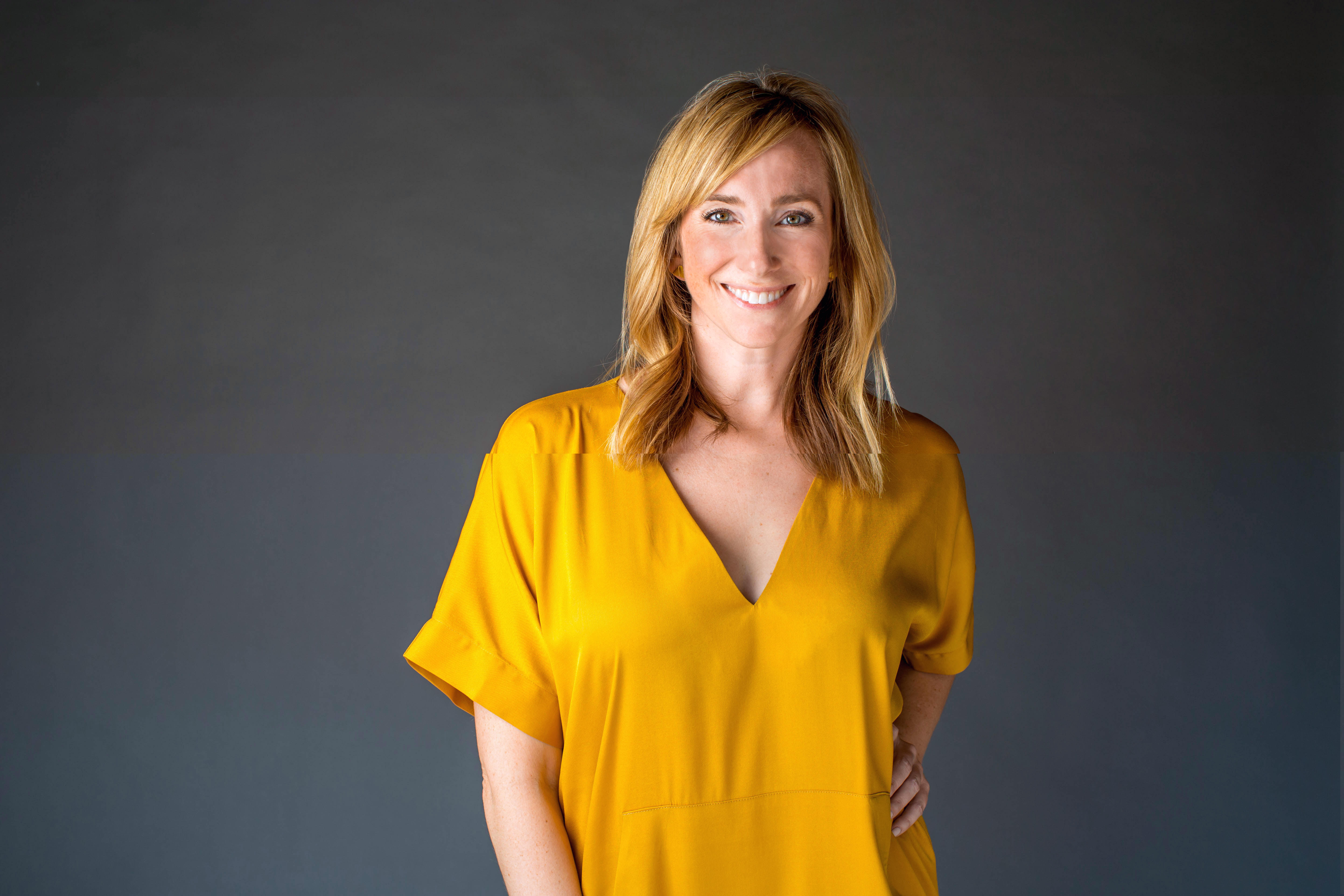 A midshot photo of Becca smiling and looking into the camera. She has mid length blonde hair and is wearing a marigold vneck dress. One hand is on her hip as the other arm hangs low.