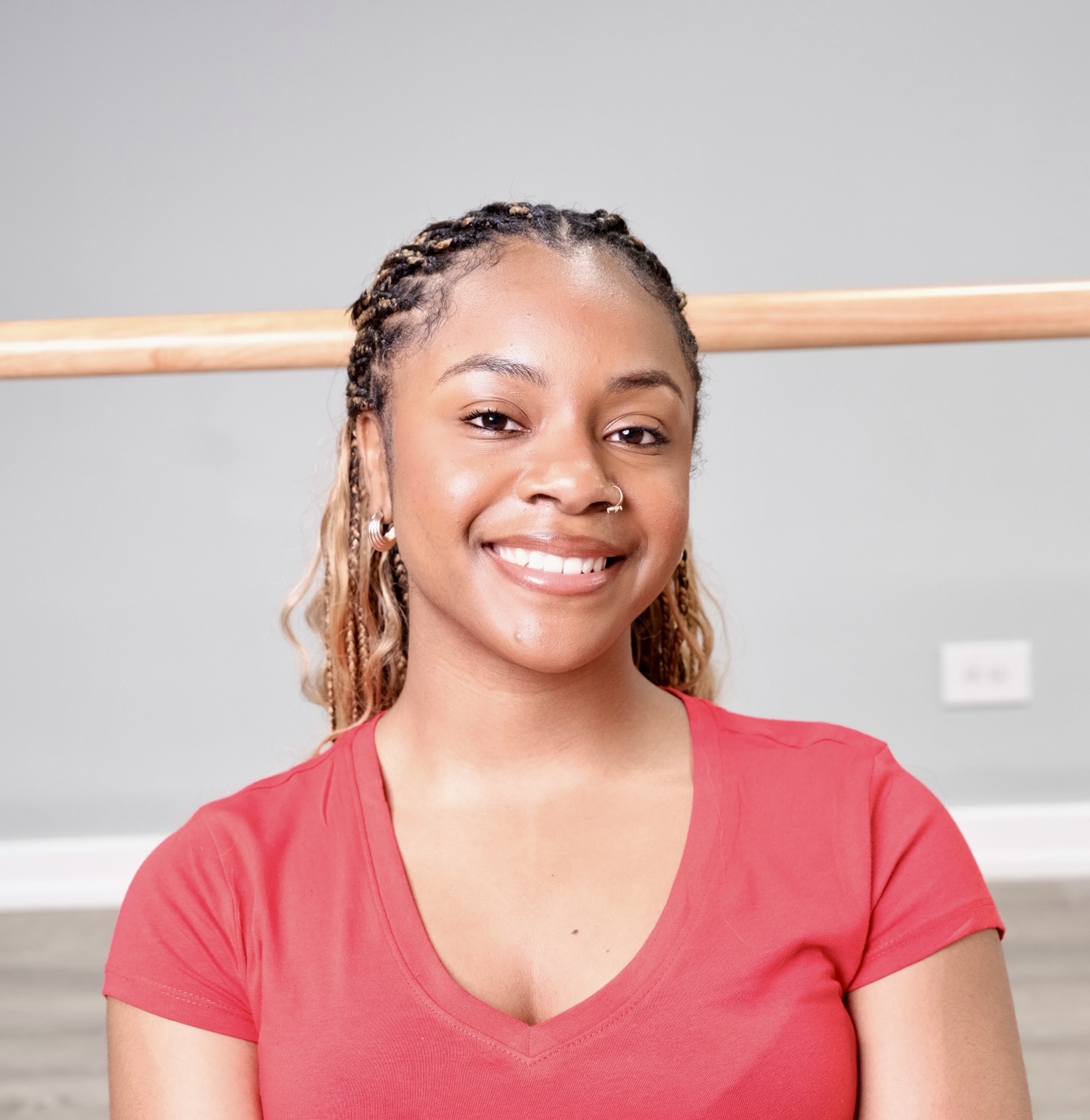 Headshot of Ashley, a smiling, African American woman with braided hair and brown eyes wearing a coral pink shirt in front of a ballet barre