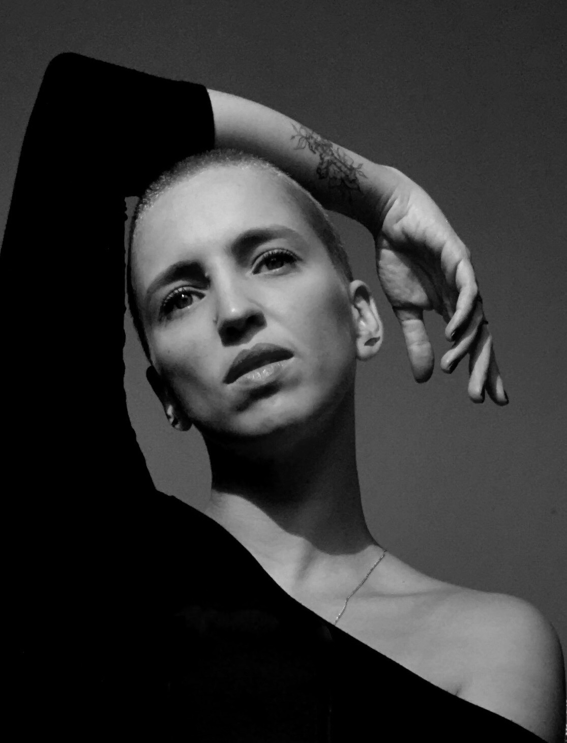 Black and white photo featuring Paula, a woman with a shaved head inclined to the left side. Her left arm is resting over her head, and she's wearing a black shirt revealing her right shoulder. There is a tattoo on her left lower arm. Paula gazes into the distance.