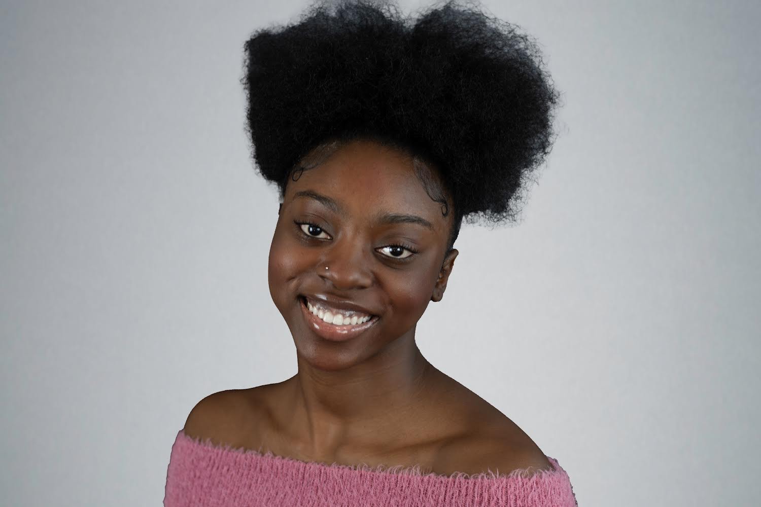 Headshot of Kenyla Jackson, a smiling young black woman with her dark hair pulled high on top of her head. She has a small nose piercing stud and is wearing an off-shoulder pink sweater