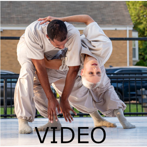 Text: Video -2 dancers in loose fitting off-white tshirts and pants are folded together on an outdoor stage