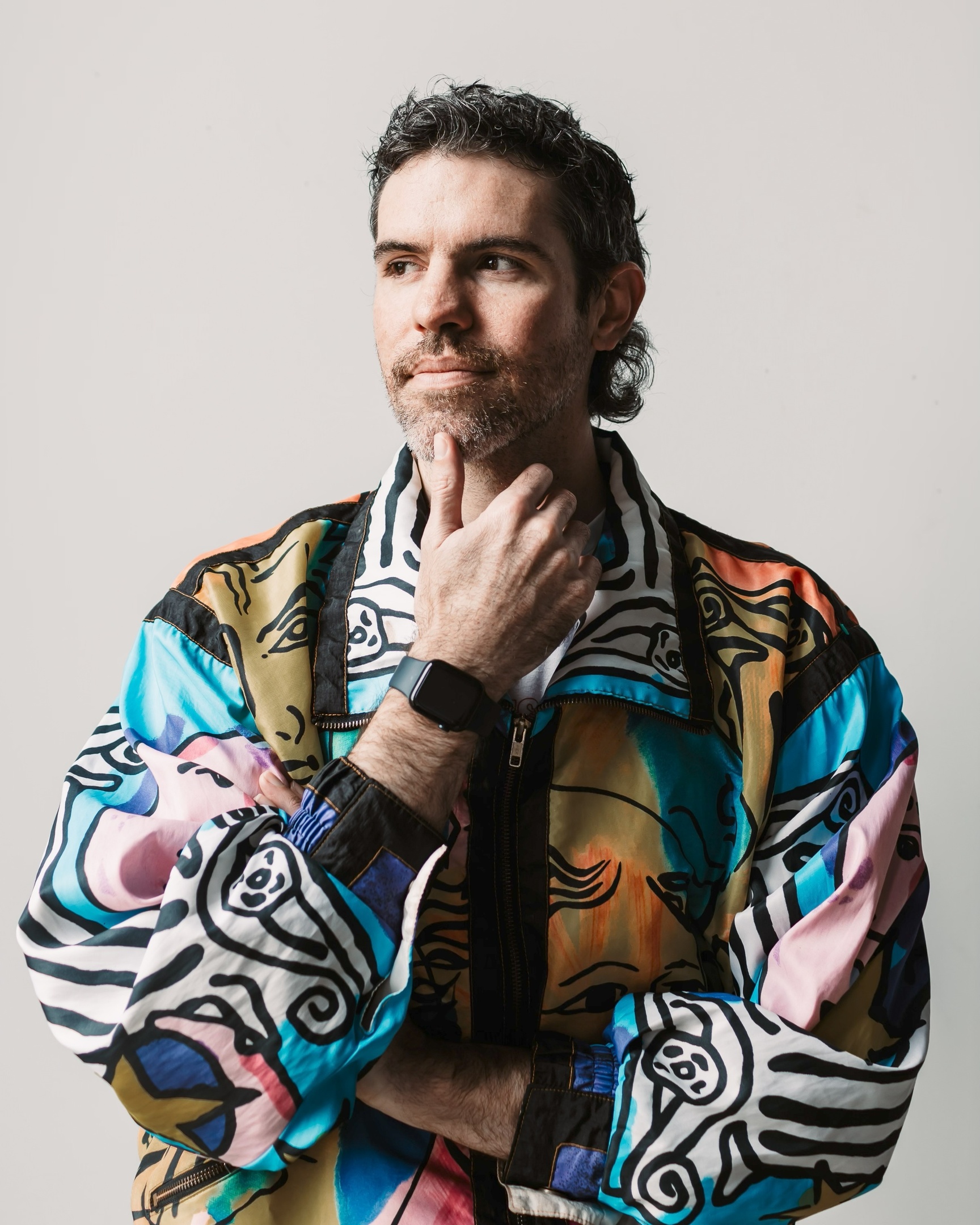 Headshot of Tom, a white man with curly dark hair, a mustache and light beard He's wearing a colorful jacket and is looking away to from the camera with his hand on his chin
