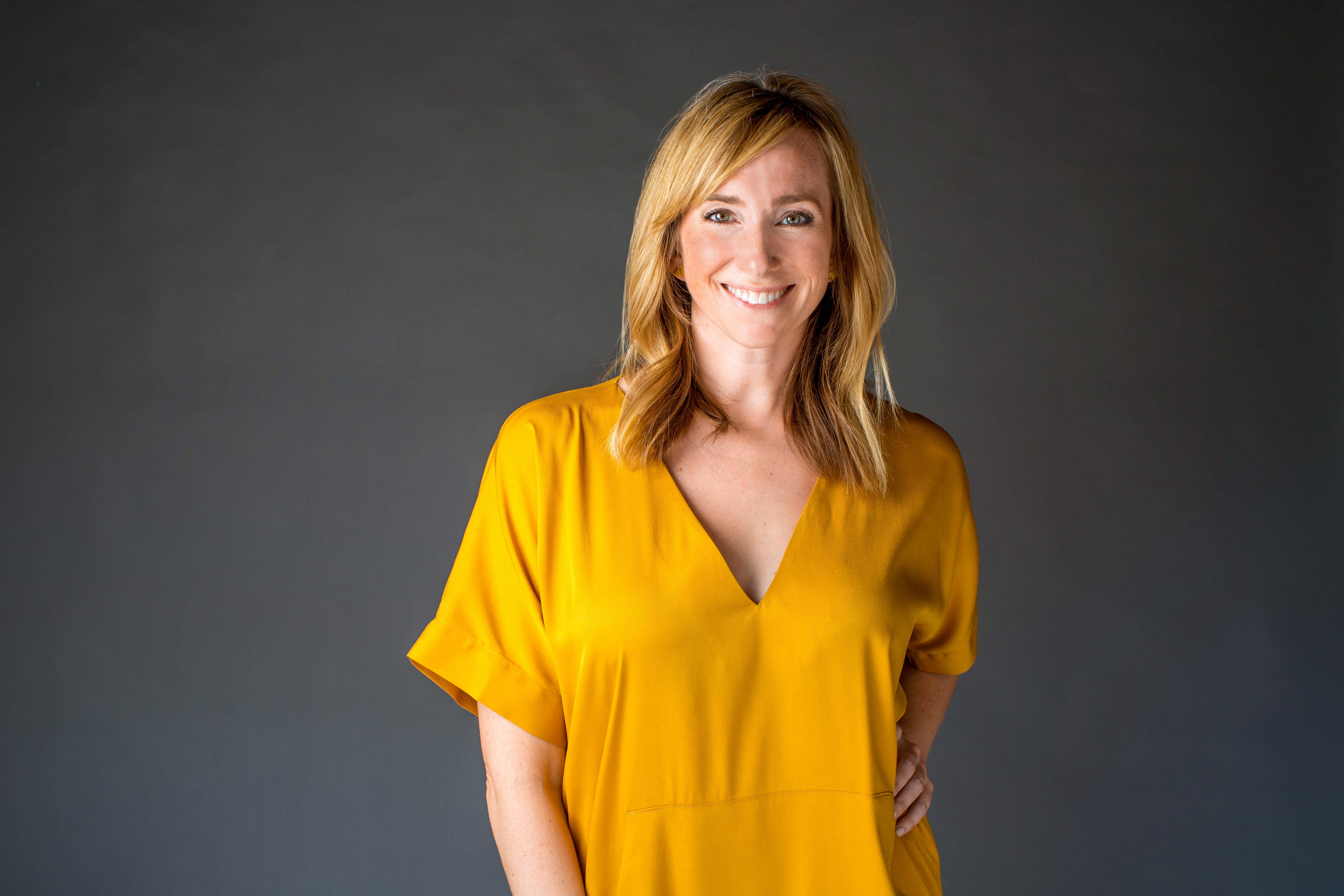 A midshot photo of Becca smiling and looking into the camera. She has mid length blonde hair and is wearing a marigold vneck dress. One hand is on her hip as the other arm hangs low.
